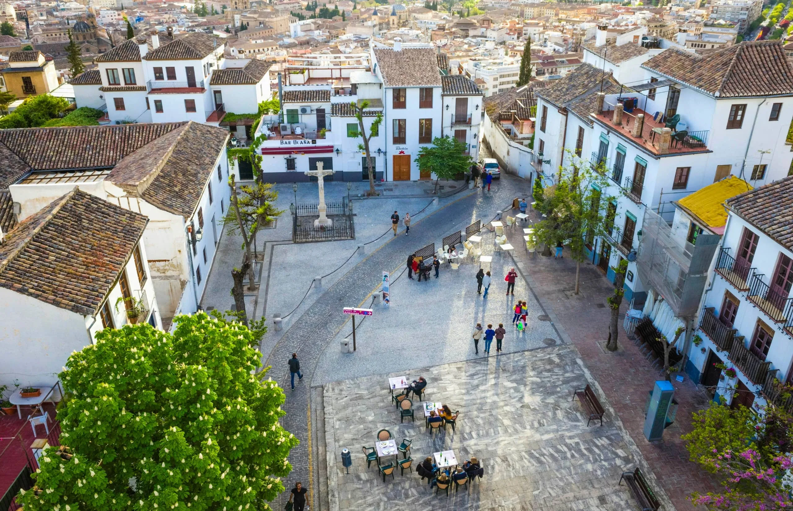 Albaicín A scenic aerial view of a plaza in the Albaicín district of Granada, Spain, featuring whitewashed buildings, cobbled streets, and outdoor cafés, with people strolling and a historic stone cross in the square.