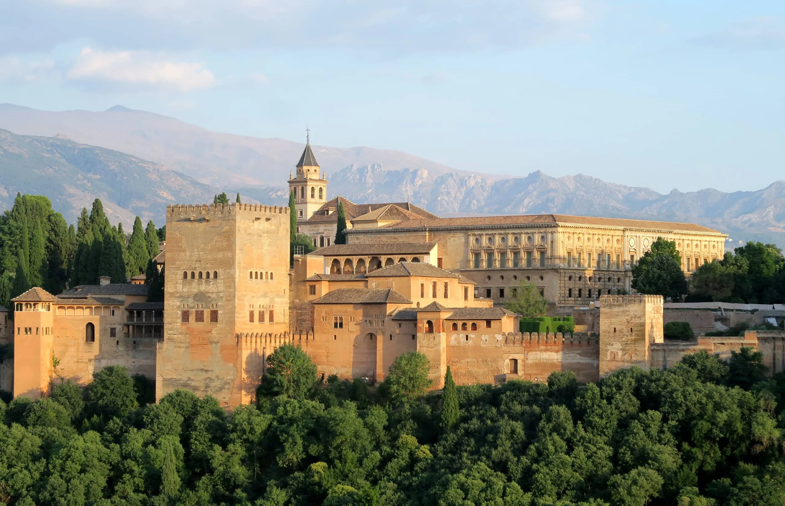 Alhambra A panoramic view of the Alhambra in Granada, Spain, with its terracotta-colored walls, fortress towers, and intricate Islamic architecture, framed by lush greenery and the Sierra Nevada mountains in the background.