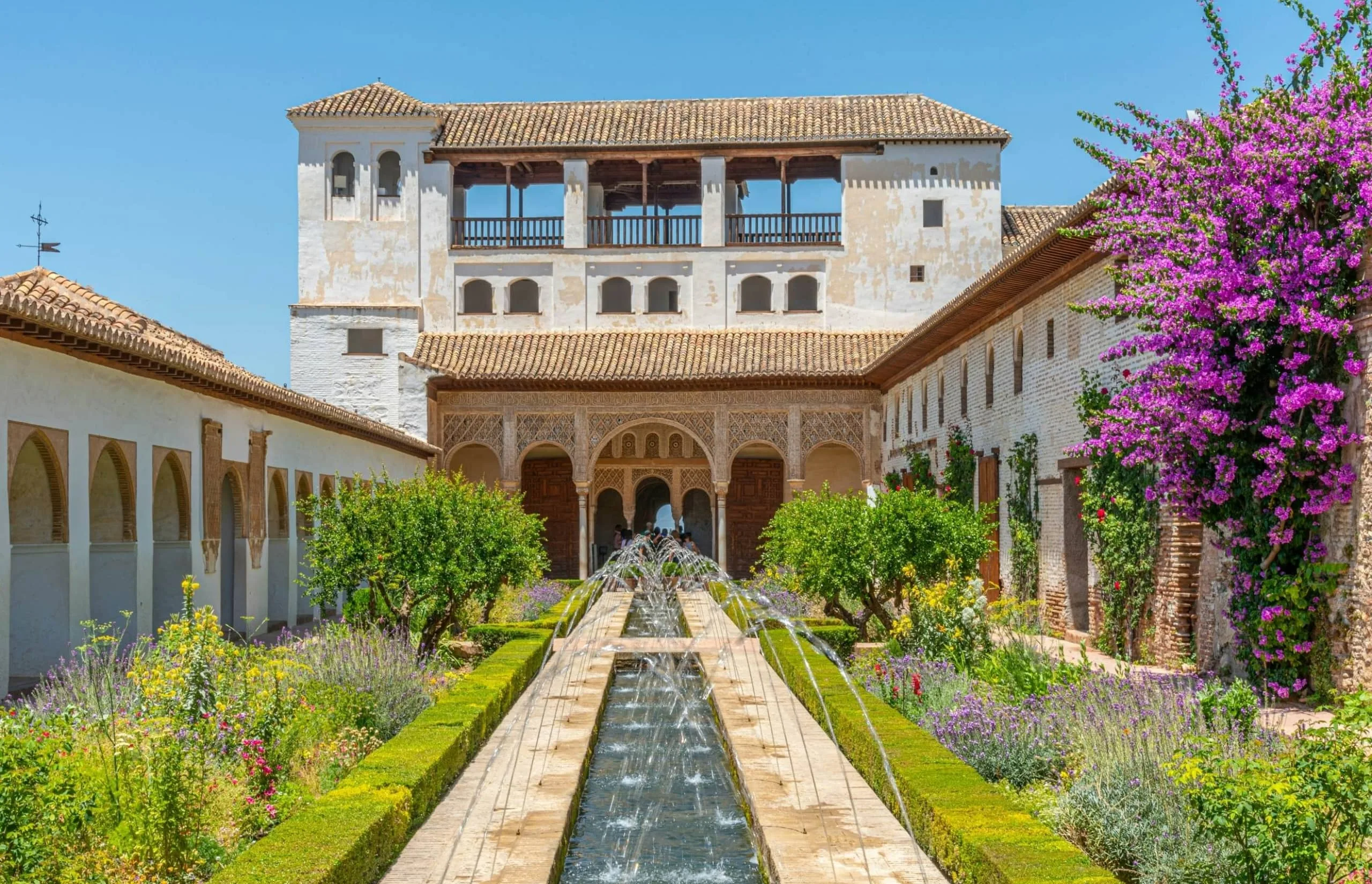 Generalife Gardens A picturesque scene of the Generalife Gardens at the Alhambra, featuring a long water canal with fountains, lush greenery, colorful flowers, and an elegant Moorish pavilion with intricate arches.