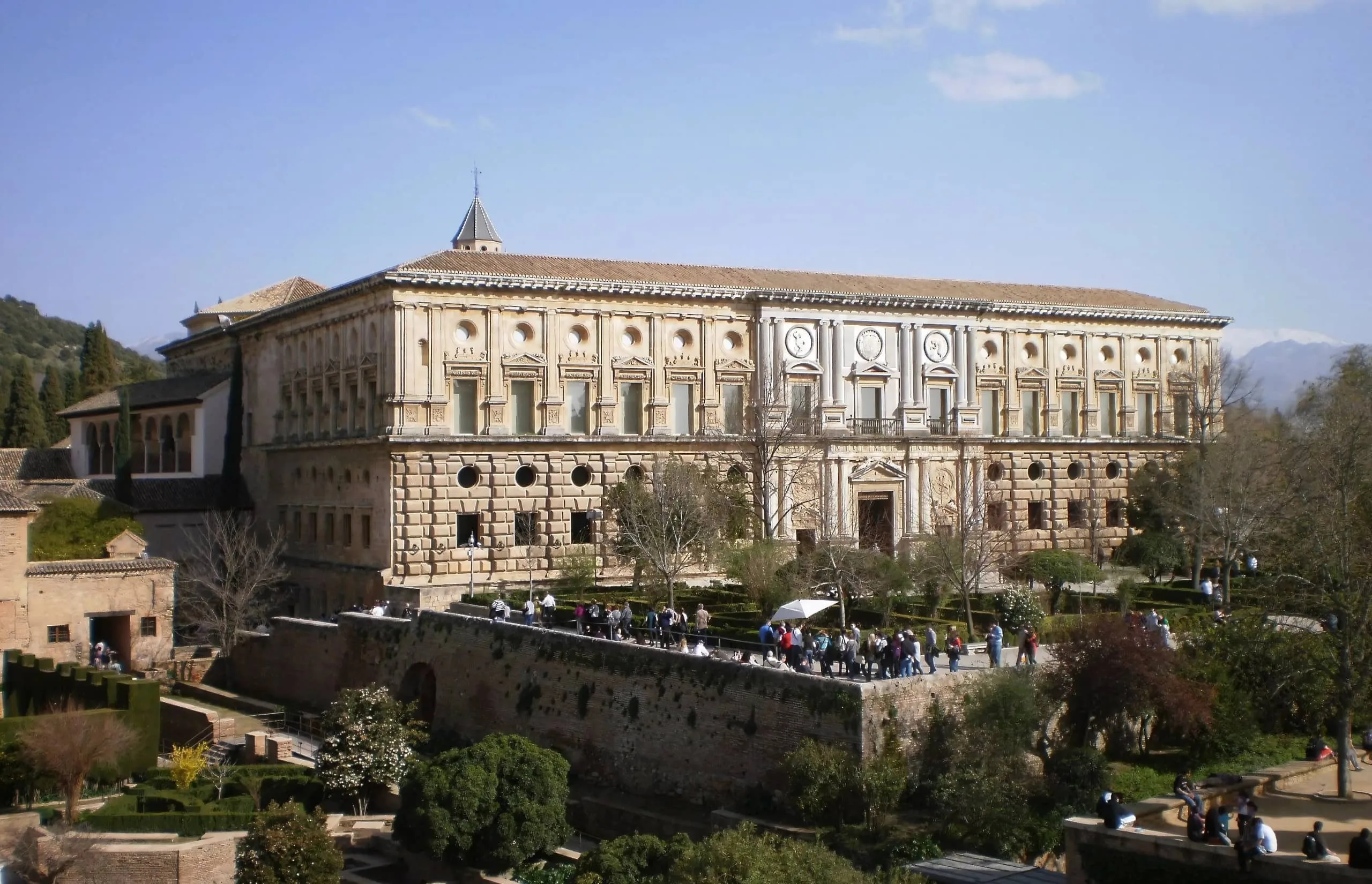 Palace of Charles V A stunning view of the Palace of Charles V in the Alhambra, Granada, featuring Renaissance architecture with an ornate façade, lush gardens, and visitors exploring the historic site under a clear blue sky.